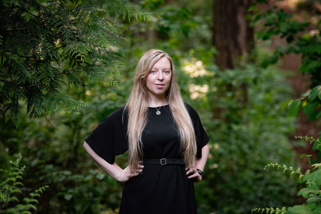 Headshot of September Herrin with waist-length blonde hair, wearing a black outfit in the middle of a green forest